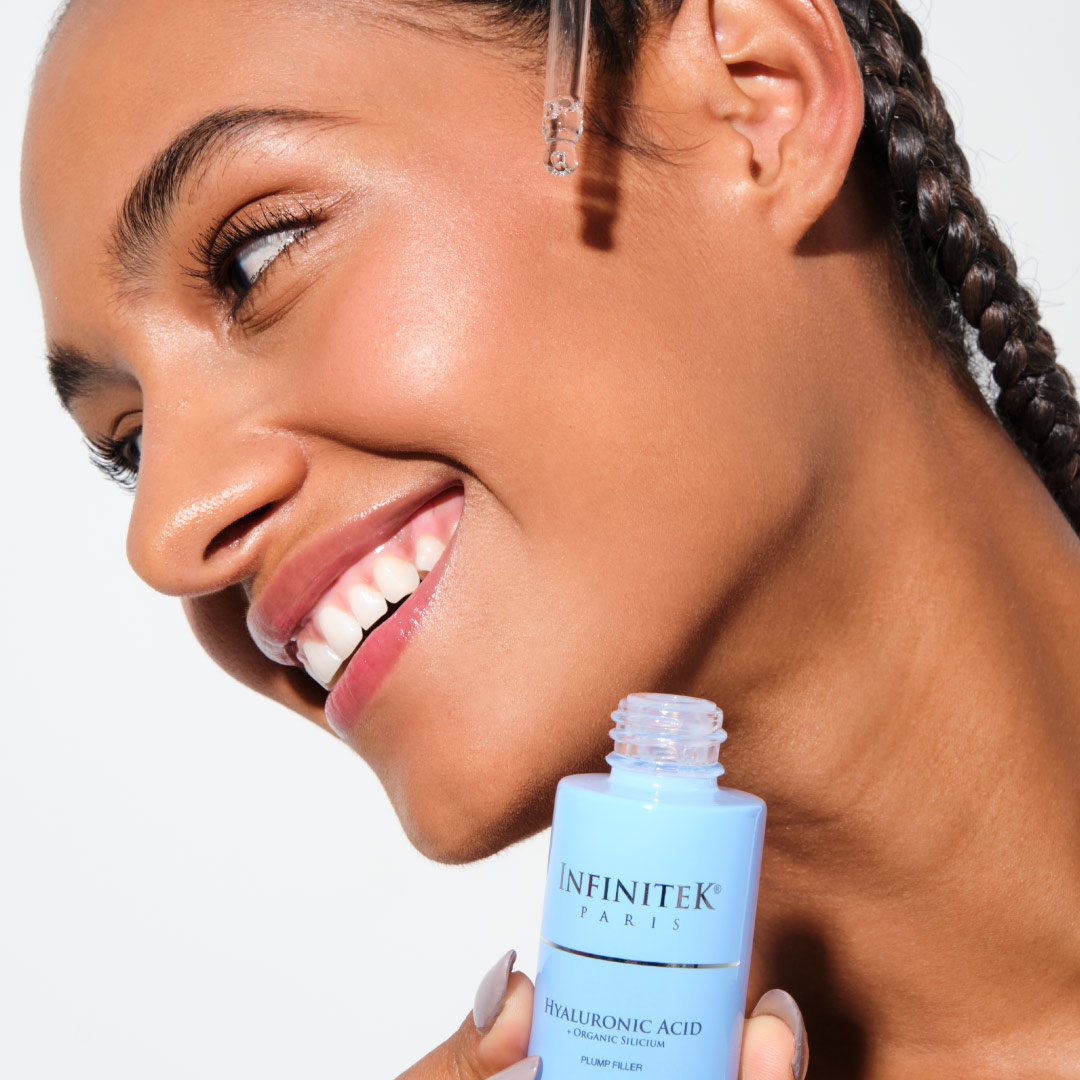 mujer modelo afro sosteniendo frasco de acido hialuronico de infinitek paris, aplicandoselo en la cara o el rostro para cuidado facial. Afro female model holding a bottle of Infinitek Paris hyaluronic acid, applying it to her face for facial care.